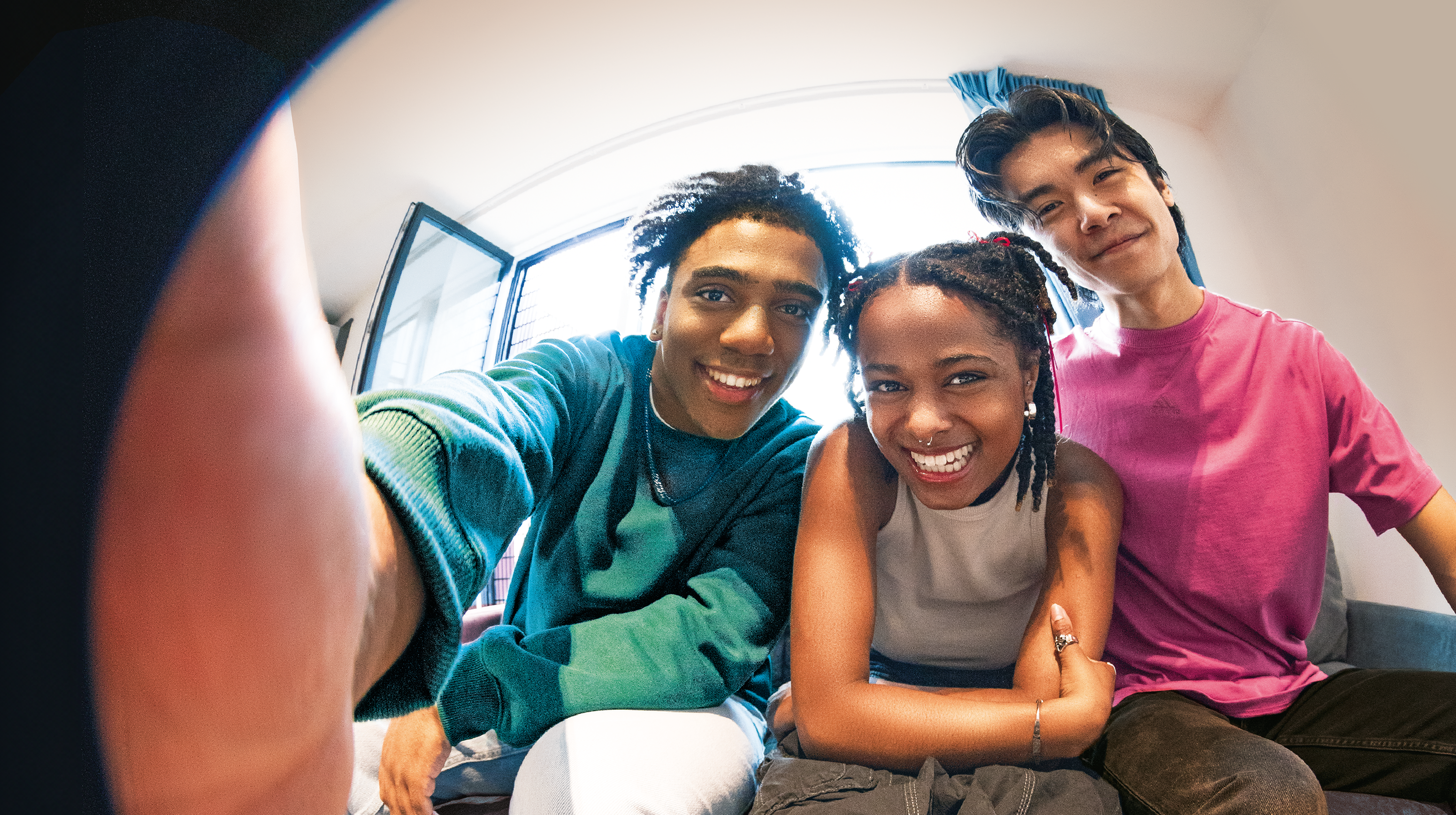 Three friends smile widely at the camera. One wears a green sweater, another a white top, and the last a pink shirt. They sit close together in a brightly lit room, capturing a cheerful and relaxed moment.